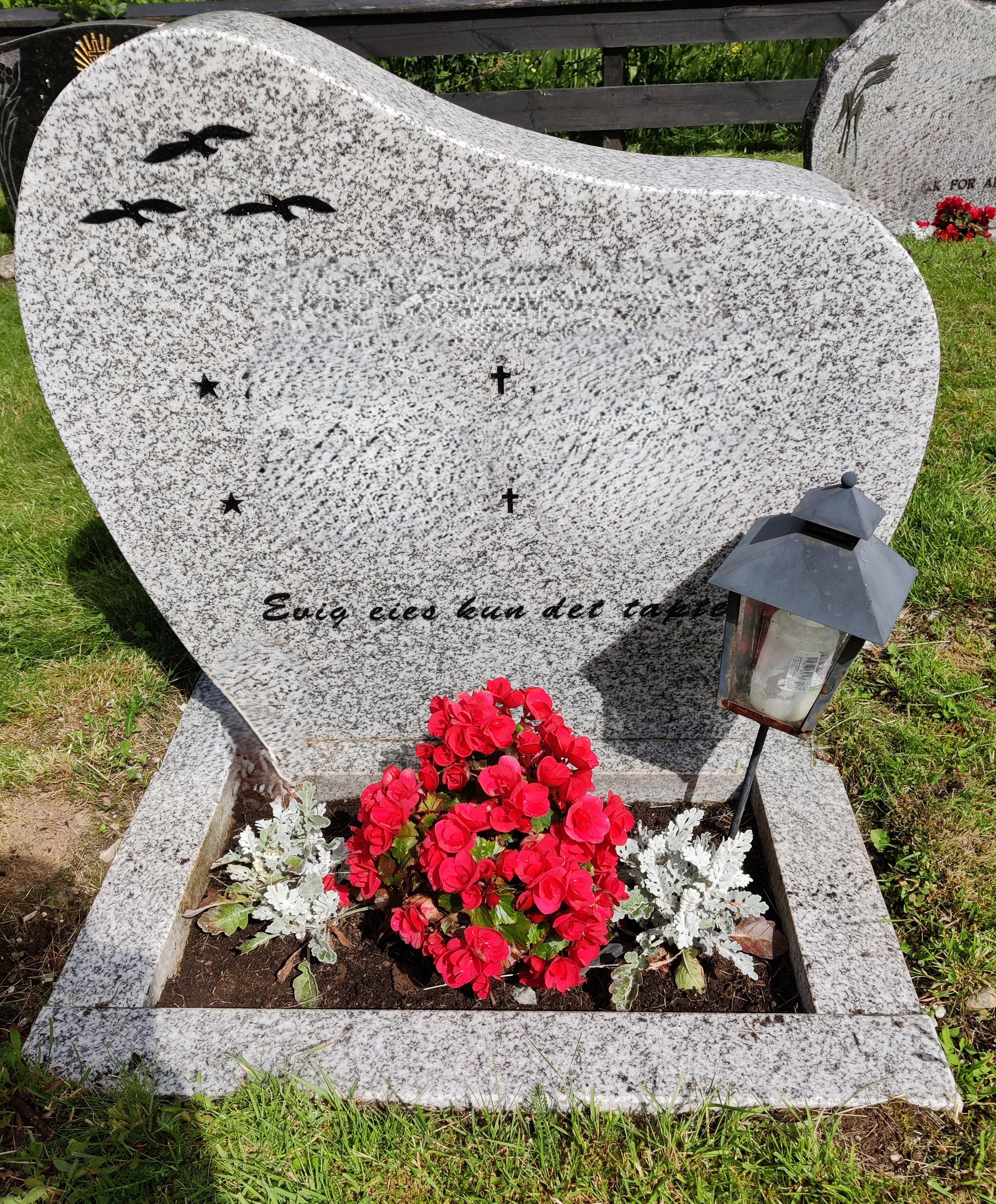 Bilete viser eit grått gravminne med tre fuglar innfelt i svart og i blomsterbeddet er det fint stellt med ein betulia og to sølvkransar.